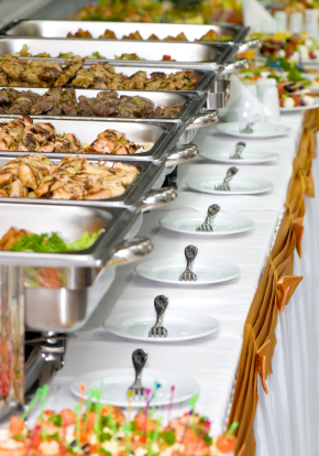 table set up with chaffing dishes filled with food and fork and plate at foot of each. Table has a tablecloth and gold decoration on the face of it.